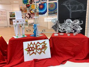 Zwei Tischgruppen mit Kunstwerken in einer Ausstellung. Auf dem linken Tisch steht eine bunte Skulptur aus verschiedenen Materialien und eine kleine weiße Struktur. Der rechte Tisch zeigt eine schwarz-weiße Zeichnung eines Tieres und mehrere kleine Kerzen. Beide Tische sind mit roten Tüchern bedeckt, und im Vordergrund ist ein auffälliges Schild mit dem Text „100 GR“ in bunten Buchstaben zu sehen. Im Hintergrund sind farbenfrohe Wandbilder zu sehen.
