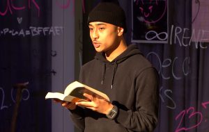 Ein junger Mann steht auf einer Bühne und liest aus einem Buch. Er trägt einen schwarzen Hoodie und eine Mütze. Im Hintergrund sind bunte Schriftzüge und Zeichnungen an einer Wand zu sehen.