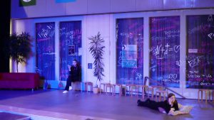 Bühne mit zwei Schauspielern in einem modernen, farbenfroh beleuchteten Raum. Im Hintergrund sind große Fenster mit bunten Graffiti und Pflanzen, während die Schauspieler in verschiedenen Positionen und Haltungen agieren. Eine Person sitzt auf einem Hocker, die andere liegt auf dem Boden und liest.