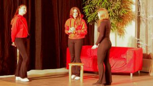 Drei Frauen stehen auf einer Bühne. Eine Frau trägt einen roten Pullover und hält einen Gegenstand in den Händen, während die anderen zwei in schwarzen Kleidung stehen. Im Hintergrund ist ein rotes Sofa und eine Pflanze zu sehen. Die Beleuchtung ist warm und die Atmosphäre wirkt lebhaft.