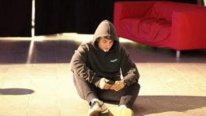 Ein junger Mensch sitzt auf dem Boden in einem Raum, trägt einen schwarzen Kapuzenpullover und schaut auf sein Smartphone. Im Hintergrund ist ein rotes Sofa zu sehen, und die Beleuchtung ist hell.