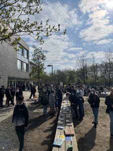 Eine Gruppe von Menschen steht im Freien vor einem modernen Gebäude. Auf einem langen Tisch sind verschiedene Gegenstände und Materialien ausgestellt. Die Szene findet an einem sonnigen Tag mit einigen Wolken statt. Viele Personen sind beschäftigt, während sie die Ausstellungsstücke betrachten und miteinander sprechen. Bäume und Grünflächen sind im Hintergrund sichtbar.