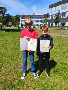 Zwei Jungen stehen auf einer grünen Wiese vor einem Gebäude. Beide halten Urkunden in den Händen. Der Junge links trägt ein rotes T-Shirt und Jeans, während der Junge rechts eine schwarze Jacke und ein dunkles Oberteil trägt. Es ist sonnig und die Umgebung wirkt freundlich.