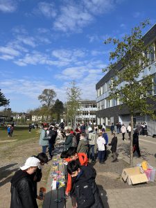 Eine Gruppe von Menschen versammelt sich im Freien vor einem modernen Gebäude. Auf einem langen Tisch sind verschiedene Gegenstände ausgestellt. Einige Personen stehen und interagieren, während andere durch den Bereich gehen. Der Himmel ist blau mit einigen Wolken, und es sind Bäume in der Umgebung zu sehen.