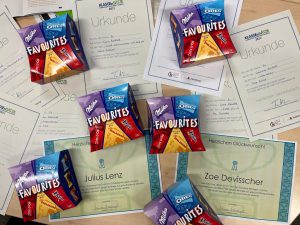 Auf dem Bild sind mehrere Urkunden und kleine Verpackungen mit Snacks angeordnet. Die Urkunden sind teilweise sichtbar beschriftet mit Namen wie 