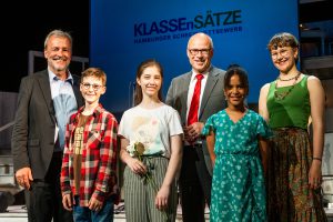 Sechs Personen stehen auf einer Bühne, lächeln und posieren für ein Foto. Im Hintergrund ist ein Banner mit der Aufschrift 