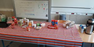 Tisch mit gestreifter Tischdecke, auf dem verschiedene Lebensmittel und Getränke stehen, darunter Kartons mit Säften, Packungen mit Snacks, eine Kanne und ein Behälter für Kaffee. Im Hintergrund ist eine Tafel zu sehen.
