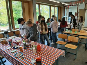 Eine Gruppe von Jugendlichen steht in einem hellen Raum mit großen Fenstern. Sie sind um einen Tisch mit einer bunten Tischdecke versammelt, auf dem verschiedene Lebensmittel und Utensilien ausgebreitet sind. Einige der Jugendlichen sind beschäftigt, während andere miteinander sprechen. Im Hintergrund sind Stühle und weitere Tische zu sehen, die den Raum ausfüllen. Es herrscht eine freundliche und aktive Atmosphäre.