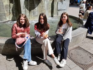 Drei Mädchen sitzen am Rand eines Brunnens in der Stadt. Sie genießen Eis und lächeln. Im Hintergrund sind einige Passanten und eine Stadtlandschaft sichtbar. Es ist ein sonniger Tag.