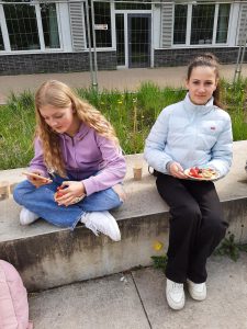 Zwei Jugendliche sitzen auf einer Betonmauer im Freien. Die linke Person hat lange, lockige Haare und trägt einen lila Pullover und Jeans. Sie hält ein Smartphone in der Hand und schaut darauf. Die rechte Person hat lange, glatte Haare und trägt eine hellblaue Jacke sowie schwarze Hose. Sie hält einen Teller mit Essen in der Hand und lächelt. Im Hintergrund sind grüne Wiesen und ein Gebäude sichtbar.