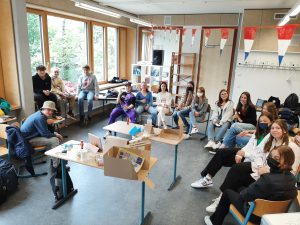 Eine Gruppe von Personen sitzt in einem Raum mit großen Fenstern, die viel Tageslicht hereinlassen. Einige Teilnehmer sitzen an Tischen, während andere auf Stühlen in einem Halbkreis um einen Tisch versammelt sind. Auf dem Tisch liegen verschiedene Materialien. Im Hintergrund sind Regale und Dekorationen zu sehen, die den Raum freundlich wirken lassen.
