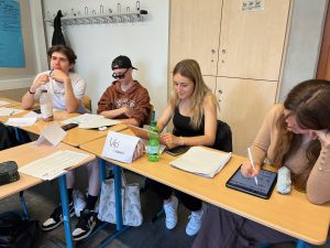 Vier Jugendliche sitzen an einem Tisch in einem Klassenzimmer. Sie sind mit verschiedenen Materialien beschäftigt, darunter Laptops und Notizen. Ein Junge trägt eine Sonnenbrille, eine andere Person hat ein Getränk vor sich. Im Hintergrund sind Möbel und eine Tafel sichtbar.