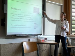 Eine Person steht vor einem Präsentationsbildschirm und zeigt auf eine Folie mit dem Titel „Textbausteine“. Die Folie enthält mehrere Punkte zu einem Thema, während die Person Notizen in der Hand hält. Im Hintergrund sind ein Tisch und eine Stuhl sichtbar.