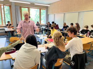 Eine Gruppe von Schülerinnen und Schülern sitzt an Tischen in einem Klassenraum. Im Vordergrund steht ein Lehrer mit blonden Haaren und einem rosa Hemd, der mit zwei Gegenständen in den Händen zu den Schülern spricht. Einige Schüler schauen aufmerksam zu, während andere mit ihren Materialien beschäftigt sind. Der Raum hat große Fenster, die viel natürliches Licht hereinlassen.