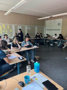 Ein Klassenzimmer mit mehreren Schülern, die an Tischen sitzen und arbeiten. Einige Schüler sind mit Laptops beschäftigt, während andere Notizen machen. An den Wänden sind Plakate und Notizen angebracht. Eine Lehrperson ist im Vordergrund zu sehen und spricht mit der Gruppe. Es herrscht eine konzentrierte Lernatmosphäre.