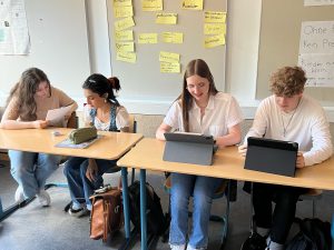Vier junge Erwachsene sitzen an Tischen in einem Klassenzimmer. Sie arbeiten an Tablets und schauen auf ihre Geräte. Im Hintergrund sind gelbe Zettel an der Wand befestigt. Die Atmosphäre wirkt konzentriert und aktiv.