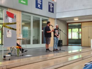 Zwei Personen stehen auf einer Bühne und sprechen in ein Mikrofon. Eine Person trägt ein schwarzes T-Shirt und kurze Hosen, die andere hat ein T-Shirt mit einem Aufdruck und eine leichte Hose an. Im Hintergrund sind Fenster und eine Wand mit verschiedenen Schildern sichtbar. Auf einem Tisch links stehen Blumen und ein Plakat. Die Umgebung wirkt hell und modern.