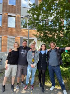 Fünf Personen stehen lächelnd in einer Reihe vor einem modernen Gebäude mit Holzverkleidung. Im Hintergrund sind grüne Bäume zu sehen. Die Gruppe besteht aus zwei Frauen und drei Männern, die verschiedene Kleidung tragen.