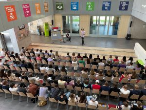 Eine große Gruppe von Menschen sitzt in einem modernen Veranstaltungsraum mit Holz- und Glas-Elementen. Im Vordergrund stehen zwei Personen auf einer kleinen Bühne, die eine Präsentation halten. Im Hintergrund sind bunte Plakate mit verschiedenen Symbolen und Texten sichtbar, die an einer Wand hängen. Der Raum hat große Fenster, die viel Tageslicht hereinlassen. Die Zuschauer sind auf Stühlen angeordnet und hören aufmerksam zu.