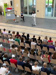 Eine Gruppe von Menschen sitzt auf Stühlen in einem großen Raum und schaut auf eine Bühne. Auf der Bühne steht eine Person mit Mikrofon, während eine andere Person auf einem Stuhl sitzt. Im Hintergrund sind große Fenster und eine Präsentationsfläche zu sehen. Die Zuschauer sind unterschiedlich gekleidet und scheinen aufmerksam zuzuhören.