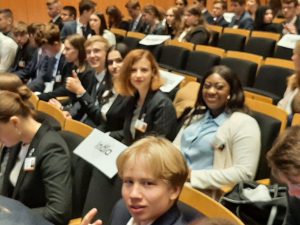 Eine Gruppe von jungen Menschen sitzt in einem Konferenzraum. Einige der Teilnehmer lächeln und zeigen mit den Händen, während sie auf Stühlen sitzen, die mit Namensschildern versehen sind. Die Anordnung der Sitze ist in Reihen, und im Hintergrund sind weitere Teilnehmer sichtbar. Alle tragen formelle Kleidung.