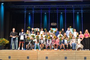 Gruppe von Jugendlichen und Erwachsenen auf einer Bühne, die Sonnenblumen halten. Die Personen stehen in mehreren Reihen, einige lächeln, während andere in die Kamera schauen. Im Hintergrund sind bunte Lichter und eine Projektionsfläche sichtbar.