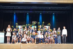 Eine Gruppe von Schülerinnen und Schülern steht auf einer Bühne, umgeben von Sonnenblumen. Im Vordergrund steht ein Mädchen mit einem Mikrofon. Die Schüler sind in unterschiedlichen Kleidungsstilen gekleidet und lächeln in die Kamera. Im Hintergrund sind bunte Lichter und Dekorationen zu sehen.