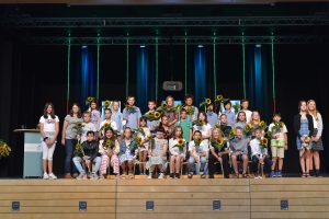 Eine Gruppe von Schülerinnen und Schülern steht auf einer Bühne. Viele halten Sonnenblumen in den Händen. Im Hintergrund sind grüne und blaue Lichtakzente zu sehen. Auf der linken Seite steht eine Lehrerin oder Betreuerin. Die Atmosphäre wirkt festlich und feierlich.