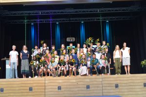 Gruppenfoto von Schülern und Lehrern auf einer Bühne. Die Schüler halten Sonnenblumen in den Händen. Im Hintergrund sind bunte Lichter und eine festliche Atmosphäre.