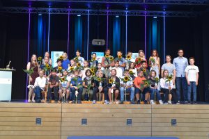 Gruppe von Schülern und Lehrern auf einer Bühne, umgeben von bunten Lichtern. Die Schüler halten Sonnenblumen und lächeln in die Kamera. Im Hintergrund sind große Bildschirme sichtbar.