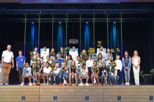 Eine Gruppe von Schülern steht auf einer Bühne, umgeben von bunten Lichtern. Sie halten Sonnenblumen in den Händen. Im Vordergrund sind mehrere Schüler in unterschiedlichen Altersgruppen und Geschlechtern zu sehen, darunter auch einige Erwachsene. Der Hintergrund zeigt eine dunkle Bühnenkulisse mit blauen und grünen Lichtern.