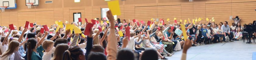 Eine große Gruppe von Menschen sitzt in einer Turnhalle. Viele halten bunte Karten in die Luft, darunter rote und gelbe. Die Atmosphäre wirkt lebhaft und engagiert, während die Teilnehmer auf einer Bankreihe sitzen und auf eine Veranstaltung oder Präsentation reagieren.