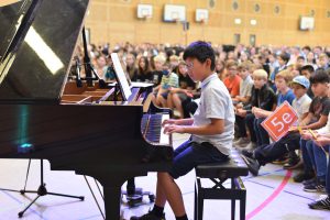 Ein junger Pianist sitzt an einem Klavier und spielt vor einem Publikum in einer Sporthalle. Im Hintergrund sind viele Zuschauer zu sehen, die aufmerksam zuschauen. Einige halten Schilder in den Händen. Die Atmosphäre ist lebhaft und konzentriert.
