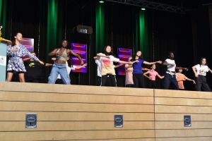 Eine Gruppe von Tänzern steht auf einer Bühne und führt synchron eine Choreografie aus. Die Tänzer sind unterschiedlich gekleidet und zeigen verschiedene Bewegungen. Im Hintergrund sind grüne Lichtstrahler und eine Leinwand sichtbar.