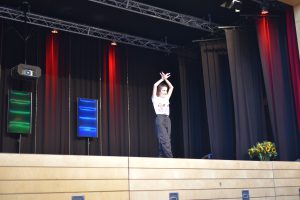 Ein Tänzer steht auf einer Bühne und hebt die Arme in die Luft. Im Hintergrund sind rote und blaue Lichter sowie Vorhänge zu sehen. Auf der rechten Seite der Bühne steht ein Topf mit Sonnenblumen.