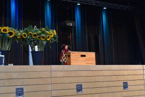 Eine Bühne mit einem Holztisch in der Mitte. Auf der linken Seite stehen mehrere Sonnenblumen in einer Vase. Im Hintergrund sind blaue und schwarze Vorhänge sowie eine Lichtinstallation sichtbar.