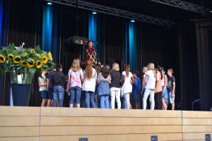 Eine Gruppe von Kindern steht auf der Bühne und schaut zu einer Person, die sie anleitet. Im Hintergrund sind Bühnenlichter und ein Blumenarrangement mit Sonnenblumen zu sehen.