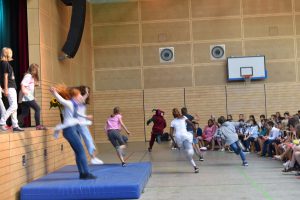 Eine Gruppe von Kindern führt eine Tanz- oder Theateraufführung in einer Turnhalle auf. Einige Kinder springen von einer Erhöhung auf eine Matte, während andere auf der Bühne stehen. Im Hintergrund sitzen Zuschauer, die die Aufführung beobachten. Die Halle hat Holzwände und eine Basketballkorb an der Wand.