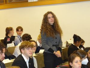 Eine Gruppe von Jugendlichen in einem Konferenzraum. Eine junge Frau mit langen, lockigen Haaren steht aufrecht und schaut aufmerksam in die Runde. Um sie herum sitzen weitere Jugendliche, die auf ihren Plätzen sitzen und auf sie blicken. Die Atmosphäre wirkt konzentriert und engagiert.