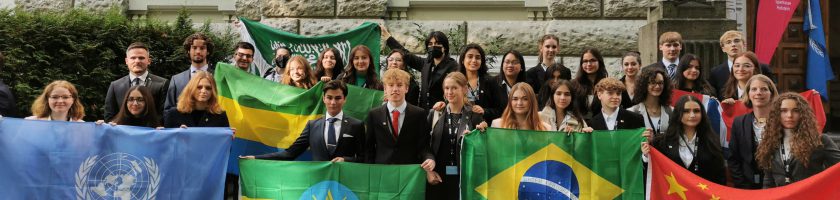 Gruppenszene mit mehreren Menschen, die an einer Veranstaltung teilnehmen. Die Teilnehmer halten verschiedene Flaggen, darunter die Flaggen von Brasilien, der Vereinten Nationen und anderen Ländern. Im Hintergrund ist ein Gebäude zu sehen. Die Personen sind in formeller Kleidung gekleidet und lächeln.