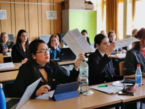 Eine Gruppe von Personen sitzt an Tischen in einem Klassenraum. Einige halten Papiere in der Hand und scheinen aktiv an einer Diskussion oder Präsentation teilzunehmen. Auf den Tischen stehen Laptops, Schreibmaterialien und Wasserflaschen. Der Raum hat große Fenster, die viel Tageslicht hereinlassen.