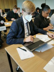 Ein junger Mann in einem Anzug trägt eine Maske und sitzt an einem Tisch mit einem Laptop. Er schreibt etwas auf einem Blatt Papier, während andere Personen im Hintergrund ebenfalls an ihren Laptops arbeiten. Die Umgebung sieht nach einem Konferenzraum oder einem Seminarraum aus.