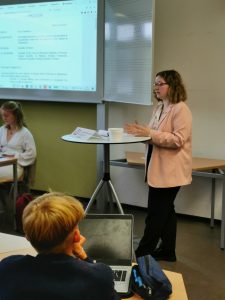 Eine Person in einem hellen Blazer steht an einem Tisch und spricht. Im Hintergrund sind weitere Personen zu sehen, die aufmerksam zuhören. An der Wand hinter der präsentierenden Person ist ein Bildschirm mit Text zu sehen. Die Umgebung wirkt wie ein klassischer Seminarraum.