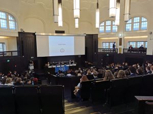 Eine große Vorlesung im Auditorium mit vielen Teilnehmern. Im Vordergrund sind mehrere Tische mit Rednern zu sehen, während auf der Leinwand eine Präsentation angezeigt wird. Das Auditorium hat hohe Decken und große Fenster, die viel Licht hereinlassen. Die Zuschauer sitzen in Reihen und schauen auf die Präsentation.