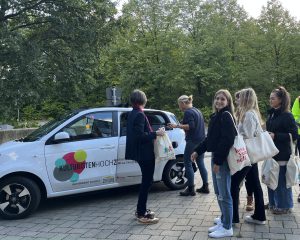 Eine Gruppe von fünf Personen steht vor einem kleinen, weißen Auto mit bunten Logos. Sie haben Einkaufstaschen in der Hand und scheinen miteinander zu sprechen. Im Hintergrund sind Bäume und eine grüne Landschaft zu sehen.