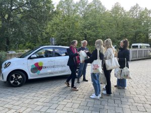 Eine Gruppe von fünf Personen steht in einem Gespräch auf einem gepflasterten Platz, neben einem weißen Auto mit buntem Logo. Im Hintergrund sind Bäume zu sehen. Die Personen tragen Taschen und scheinen an einem Austausch interessiert zu sein.