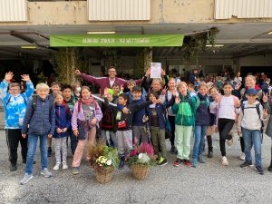 Eine Gruppe von Kindern und Jugendlichen steht mit fröhlichen Gesichtsausdrücken und erhobenen Händen vor einem Gebäude. Sie halten Blumensträuße in Körben und posieren für das Foto. Im Hintergrund ist ein grünes Banner mit der Aufschrift „AMBIENTE TRAUM – TEATRO“ zu sehen.