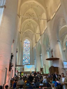 Blick in das Innere einer Kirche mit hohen, gewölbten Decken und einem großen, bunten Fenster. Viele Menschen sitzen in Reihen, während einige auf eine Leinwand schauen. Der Raum ist hell erleuchtet, und es gibt verschiedene religiöse Statuen und Ausstattungen.