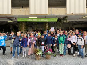 Eine Gruppe von Kindern und Jugendlichen steht lächelnd vor einem Gebäudeeingang, umgeben von Pflanzenarrangements. Im Hintergrund ist ein Banner mit der Aufschrift „Allerlei rund um die Natur“ zu sehen. Die Kinder tragen unterschiedliche Kleidung und einige halten Blumen oder andere Gegenstände in der Hand.