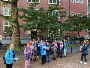 Gruppe von Kindern und Erwachsenen steht im Freien vor einem Gebäude, umgeben von Bäumen. Die Kinder tragen verschiedene bunte Jacken und halten etwas in den Händen. Die Szene wirkt fröhlich und lebhaft.